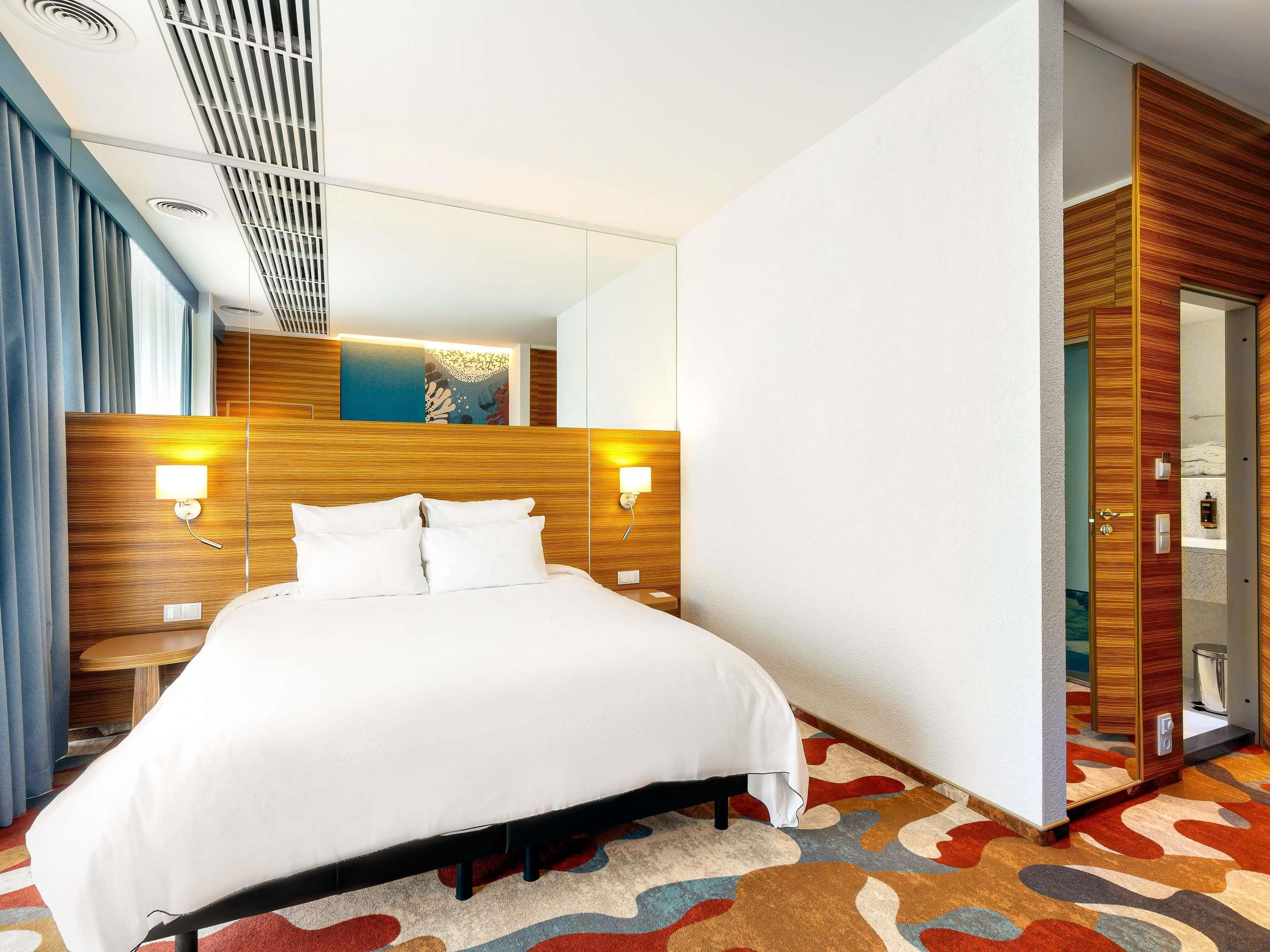 A hotel room with a bed, bedside lamps, a headboard, a mirror, a shower, and a patterned carpet.