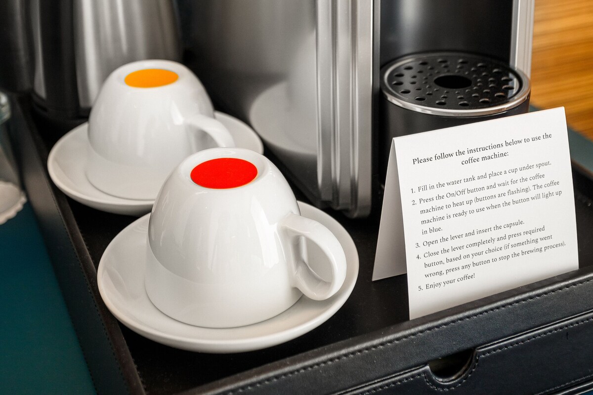 A coffee machine with instructions on a tray, two white cups with red lids, and a silver kettle.