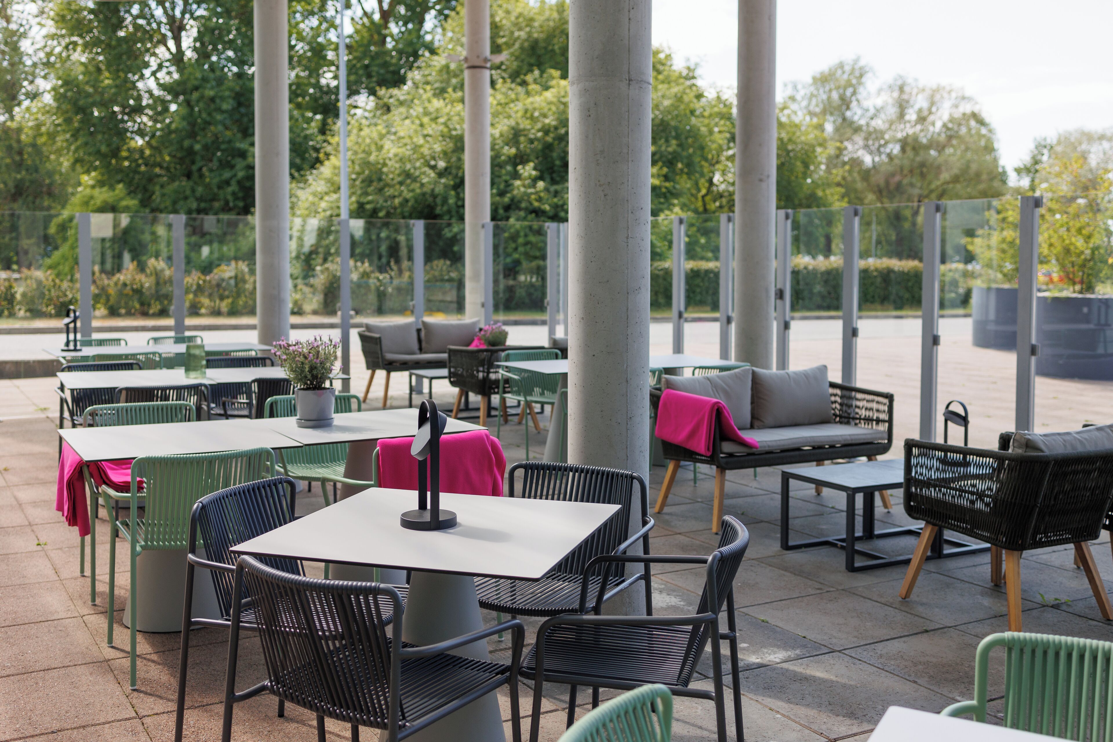 En udendørs terrasse med borde og stole, omgivet af høje søjler og glasgelændere.