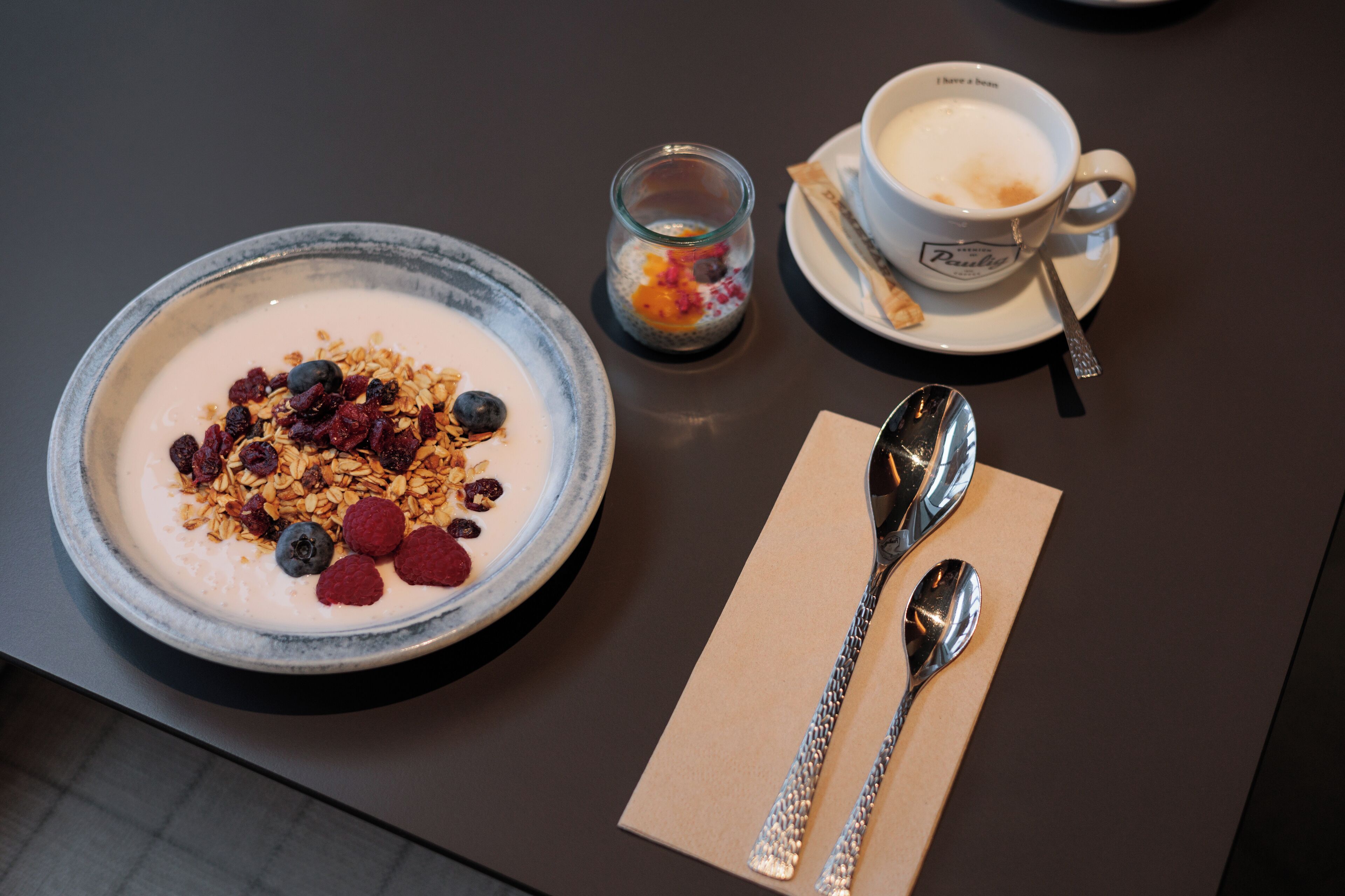 Ein Frühstückstisch mit einer Schüssel Joghurt, belegt mit Granola und Beeren, einer Kaffeetasse und einem kleinen Glas gemischten Obst.