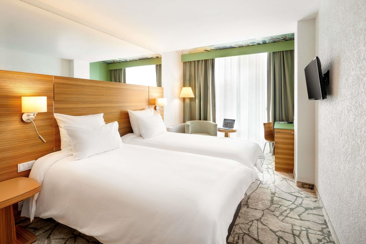 A hotel room with two beds, a desk, a chair, a TV mounted on the wall, and a window with curtains.