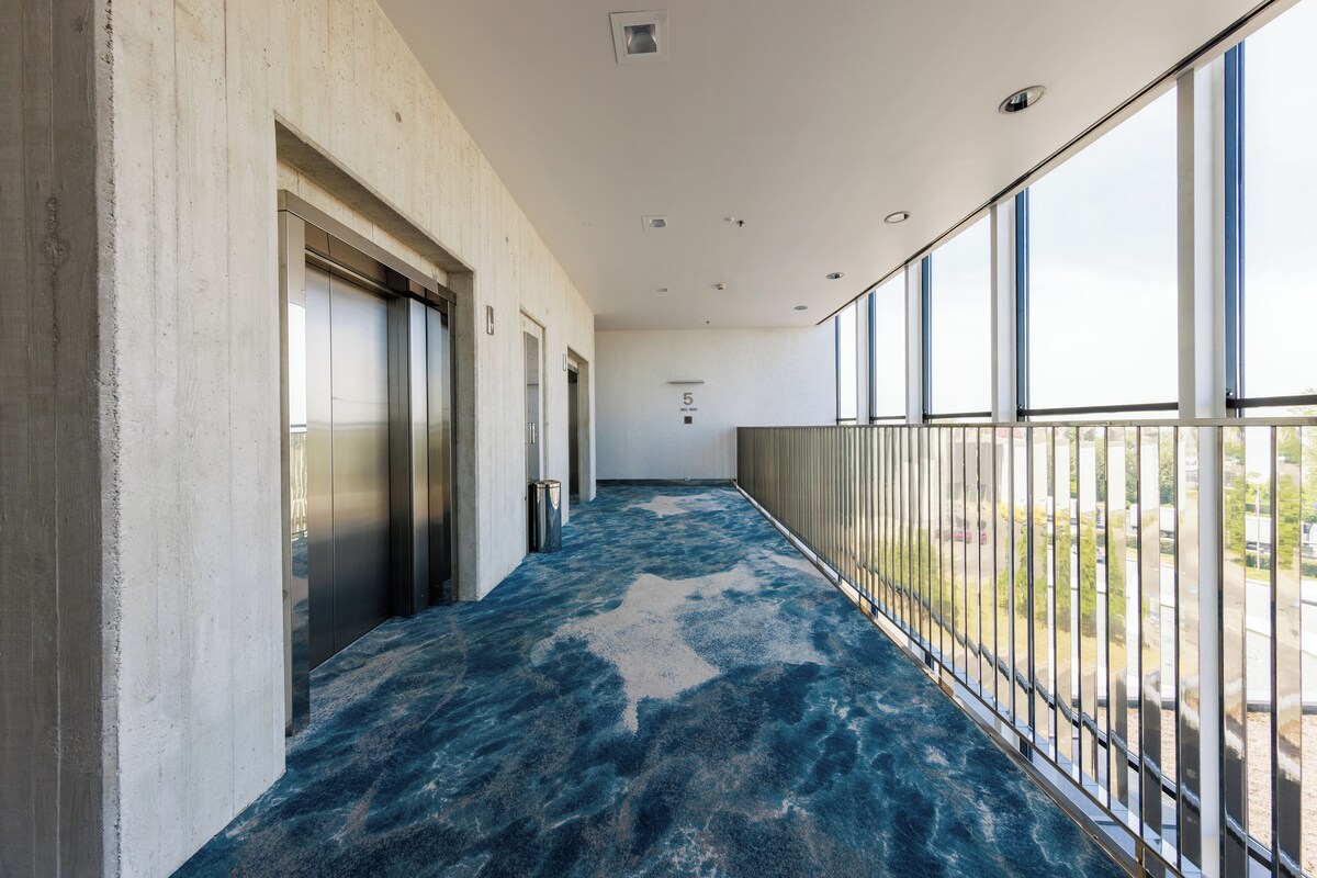 A corridor with a blue patterned floor, elevators, and large windows offering a view of the outdoors.