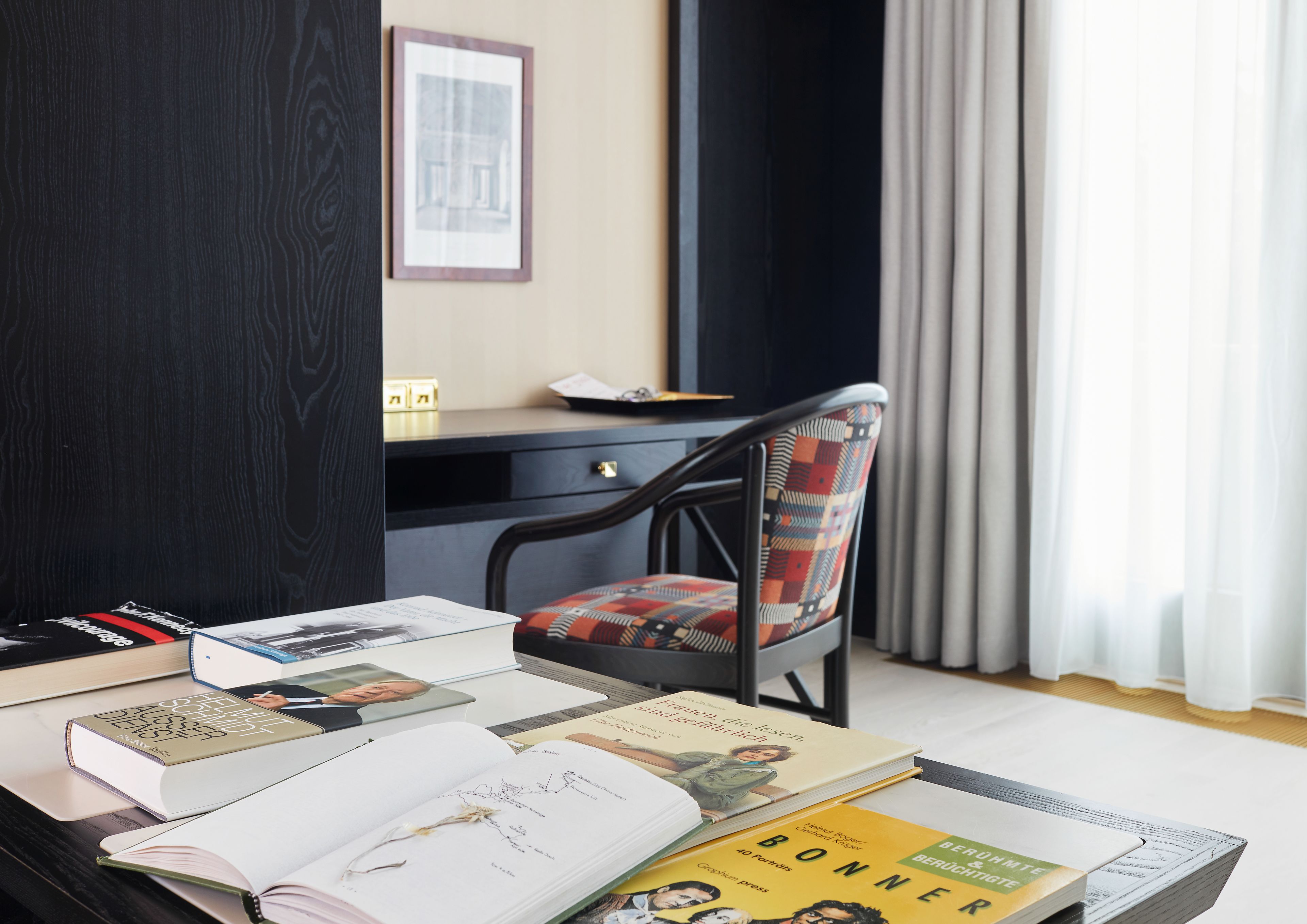 Ein Zimmer mit Schreibtisch, Stuhl und Bett. Auf dem Schreibtisch liegen Bücher und im Eck steht ein Bett.