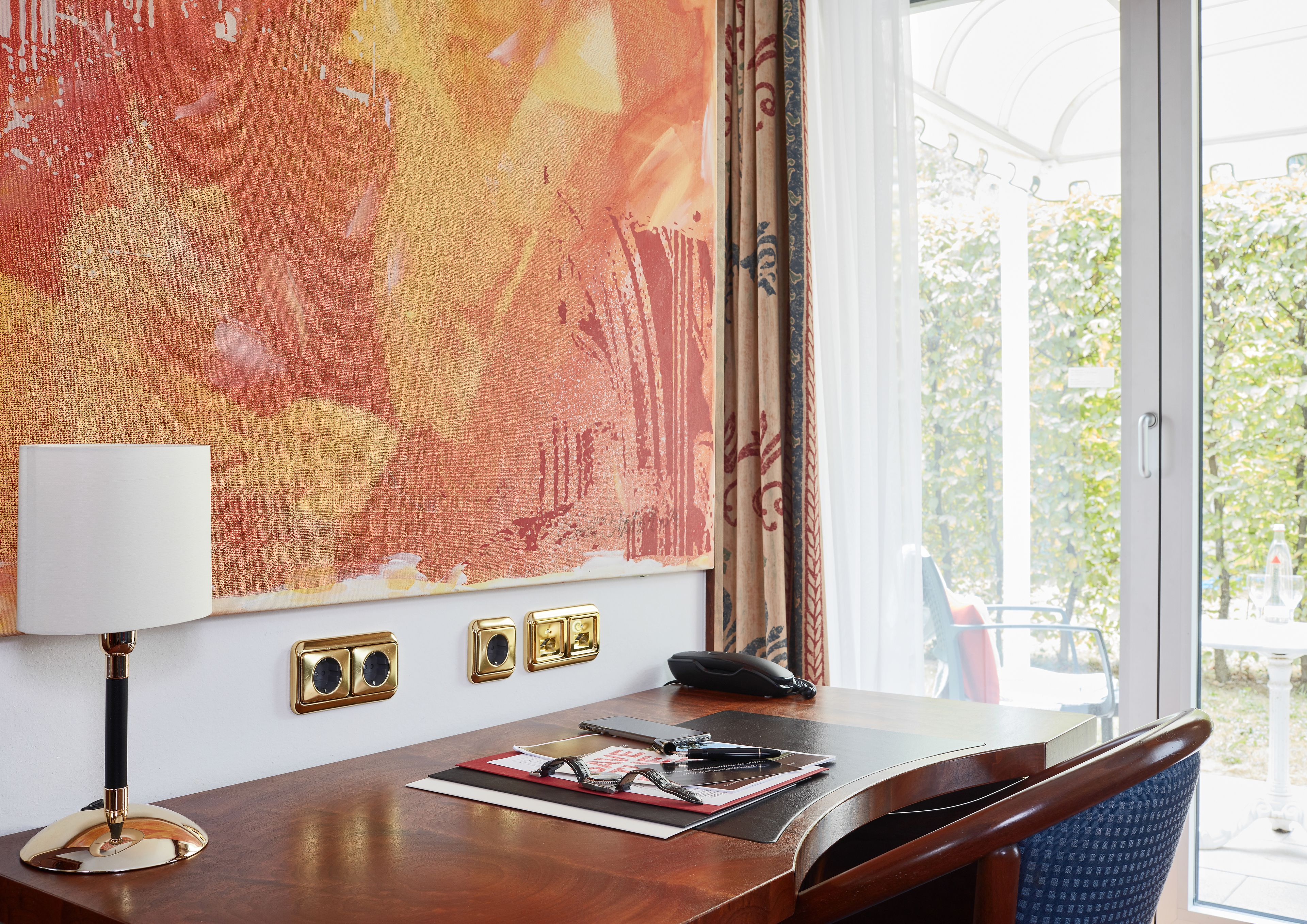 A hotel room with a large abstract painting on the wall, a desk with a lamp, phone, and some personal items, and a view of greenery outside.