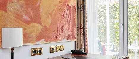 A hotel room with a large abstract painting on the wall, a desk with a lamp, phone, and some personal items, and a view of greenery outside.