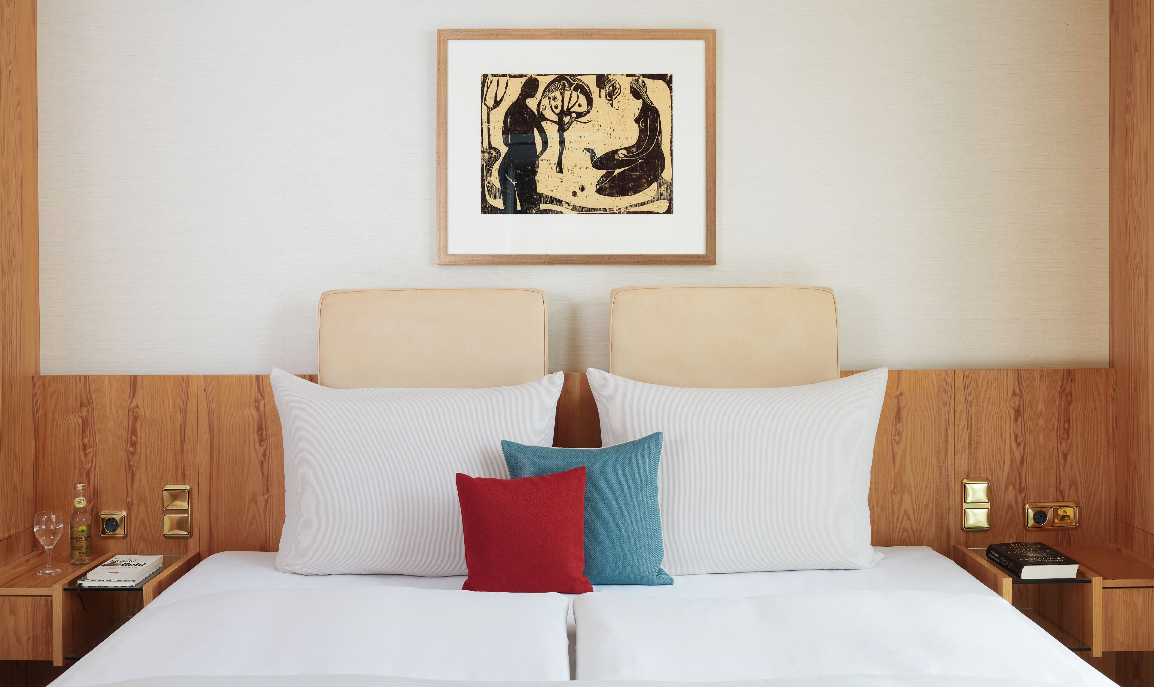 Una habitación de hotel con cama, almohadas, una mesita de noche con un libro, una botella y un vaso.