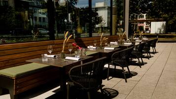 Een buiten eetgedeelte met houten banken en stoelen, een lange tafel gedekt voor een maaltijd, en een uitzicht op de straat en gebouwen op de achtergrond.