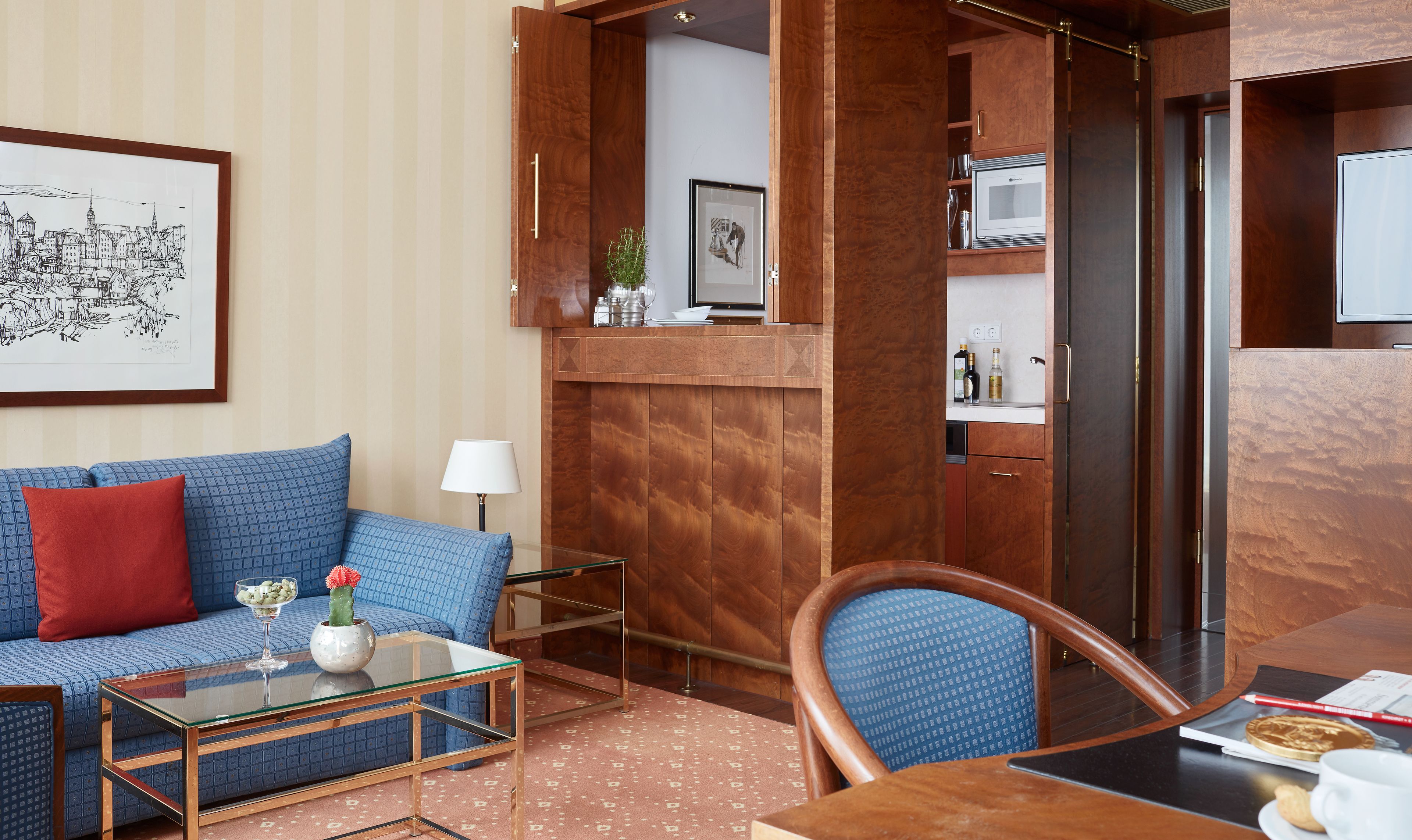 Una moderna camera d'hotel con un divano blu a fantasia, un tavolino in vetro, una scrivania in legno e un tavolo da pranzo. Si nota un'opera d'arte raffigurante un panorama urbano appesa alla parete, una piccola pianta e un forno a microonde nella zona cucina.