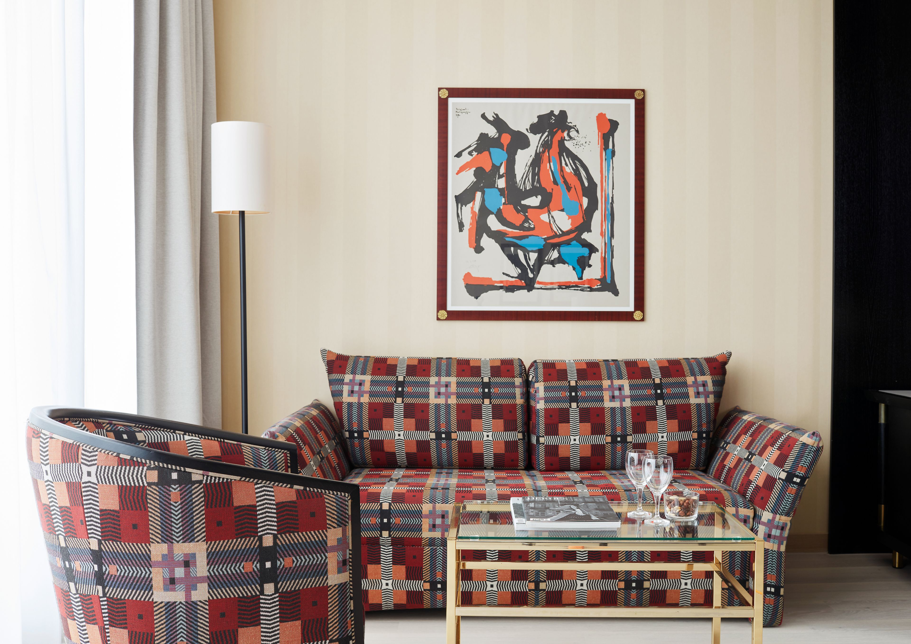 A living room with a patterned sofa, a glass coffee table, and a framed abstract painting on the wall.