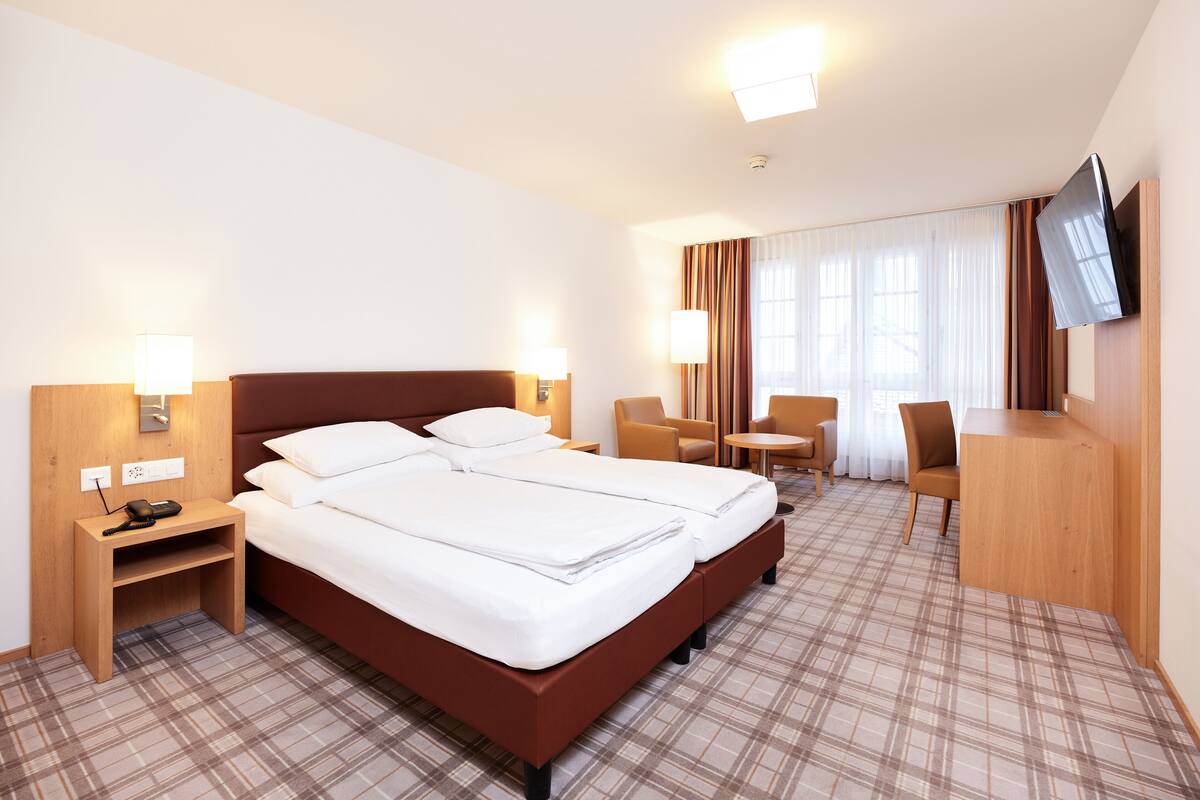 Quarto de hotel com cama de casal, escrivaninha, cadeira, televisão e mesinha.