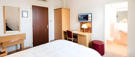 A hotel room with a bed, a desk, a chair, a red ottoman, a mirror, and a TV.