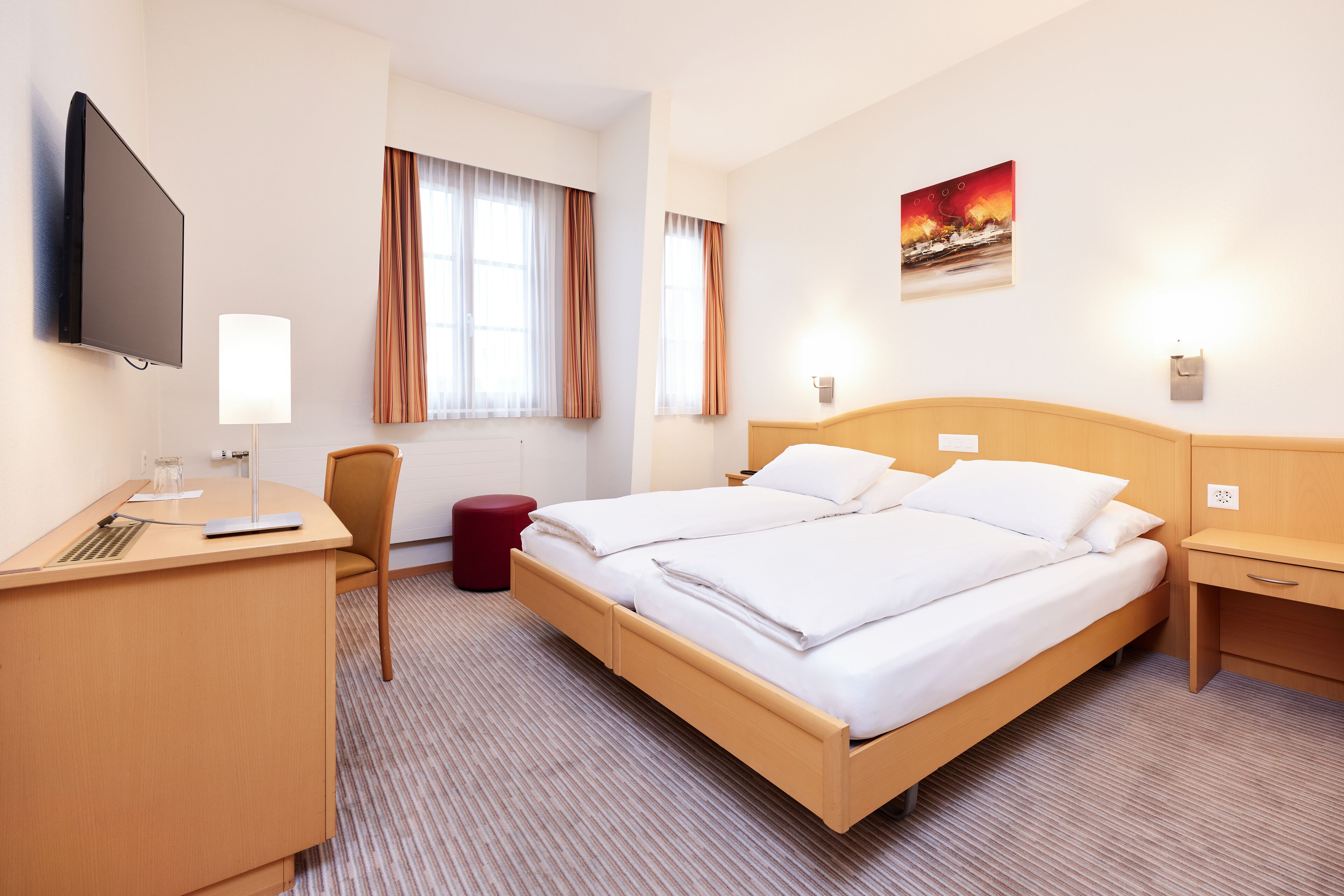 A hotel room with a bed, a desk, a chair, a TV, and a painting on the wall.