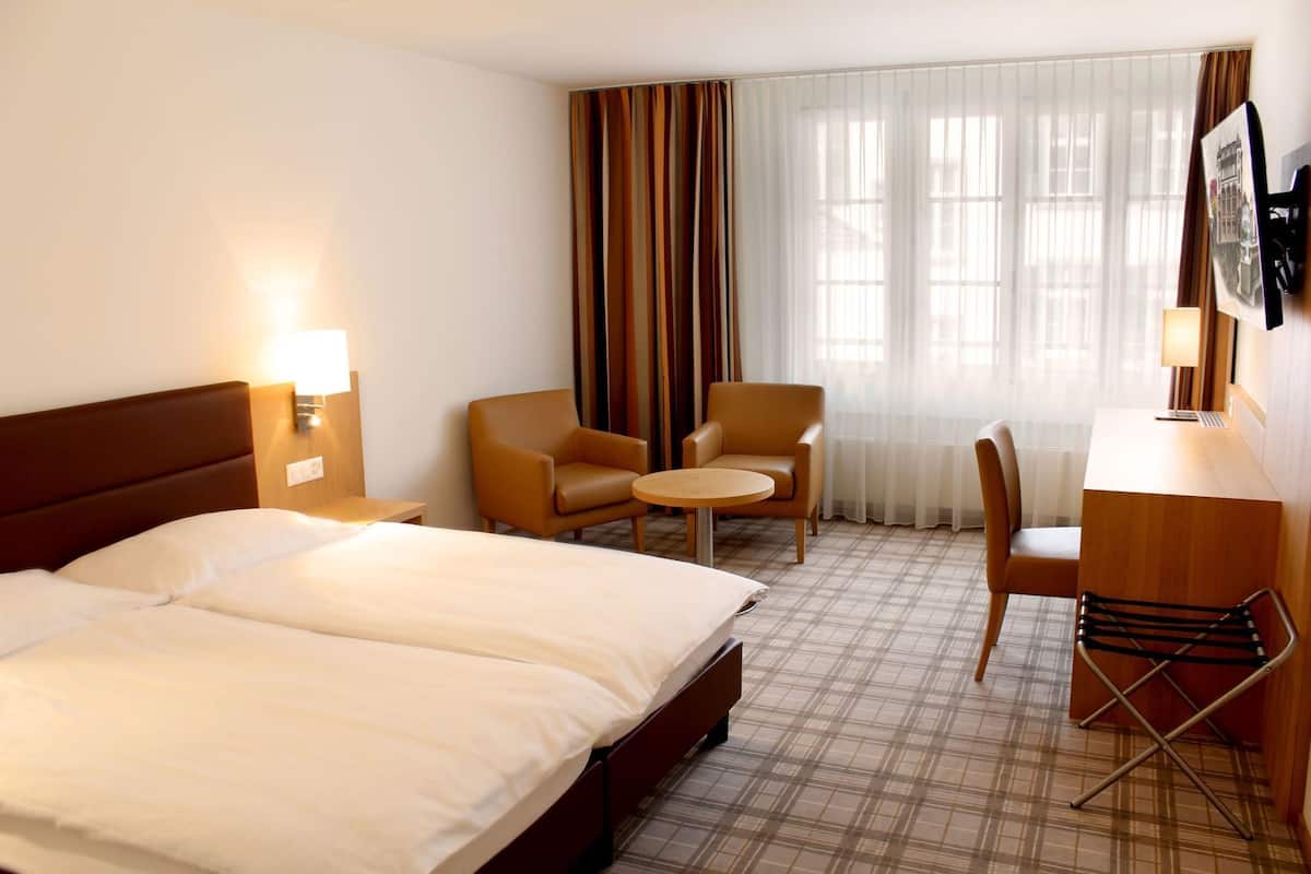 Quarto de hotel com uma cama de casal, duas poltronas, uma mesa redonda pequena, uma escrivaninha com cadeira e uma janela grande com cortinas.