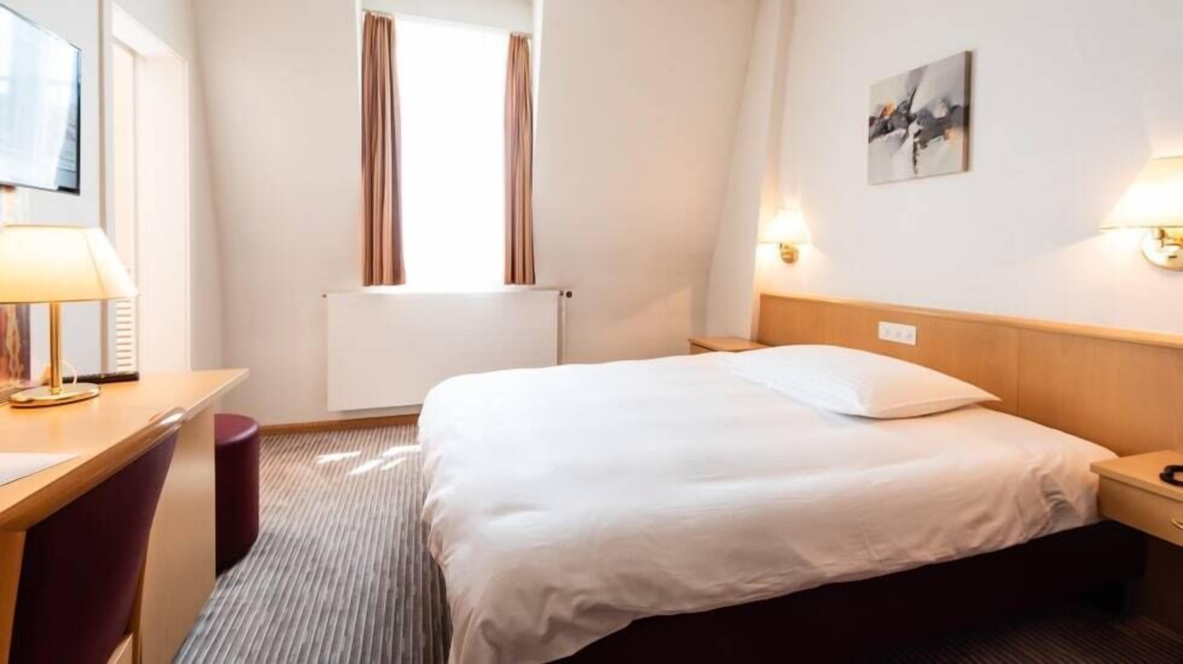 A hotel room with a bed, a desk, a chair, a window with curtains, a radiator, and a wall-mounted lamp.