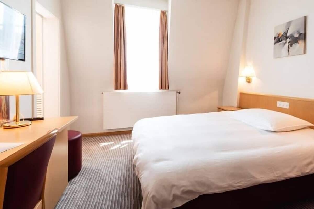 Quarto de hotel com cama, escrivaninha, cadeira, janela com cortinas, radiador e luminária na parede.