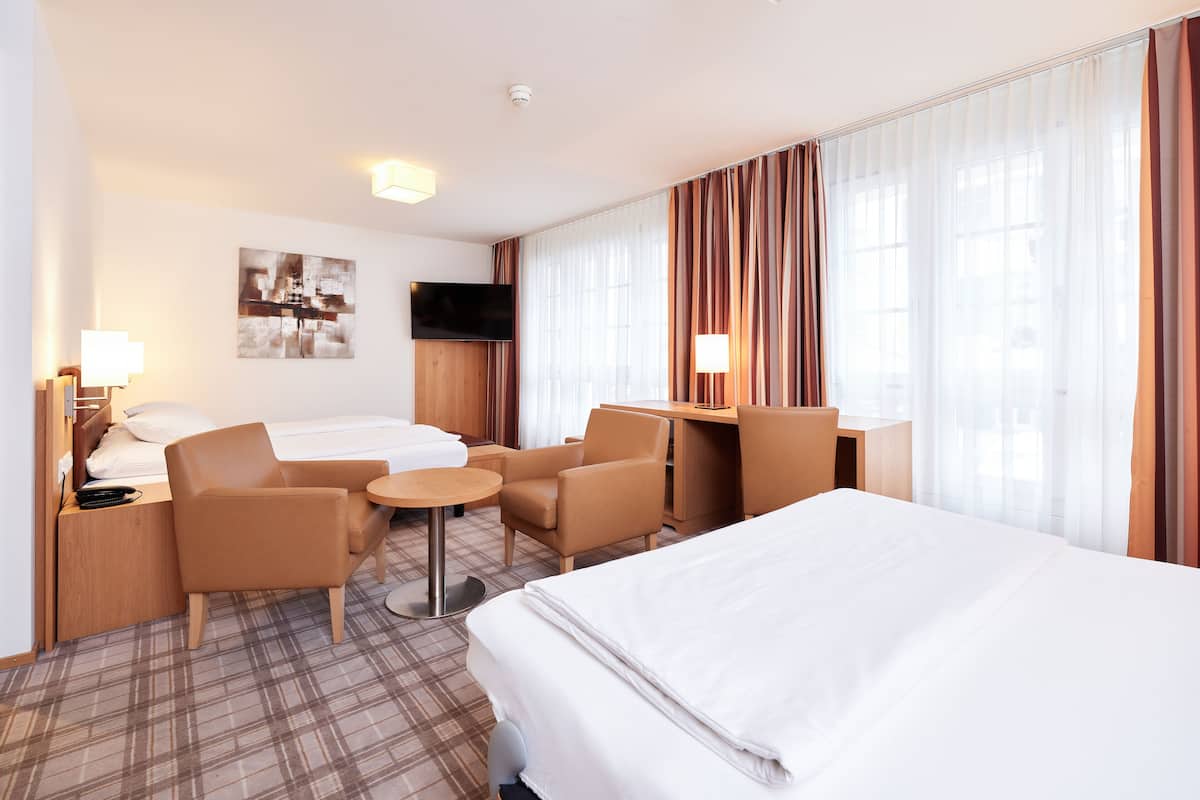 Quarto de hotel com cama, duas poltronas, uma mesinha redonda, escrivaninha, televisão e uma janela grande com cortinas.