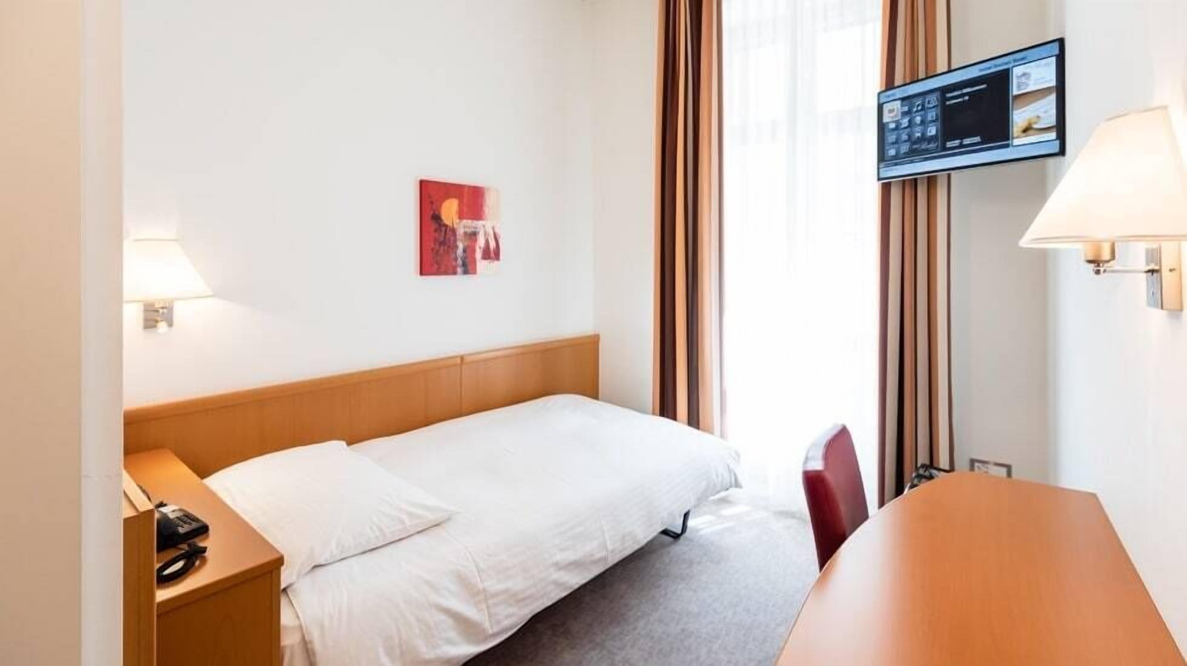 A hotel room with a bed, a desk, a chair, a TV mounted on the wall, and a window with curtains.
