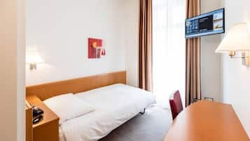 A hotel room with a bed, a desk, a chair, a TV mounted on the wall, and a window with curtains.