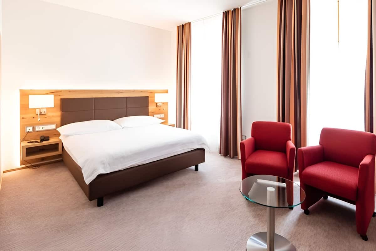 Quarto de hotel com cama, duas cadeiras vermelhas, mesa de vidro e mesa de cabeceira com abajur.