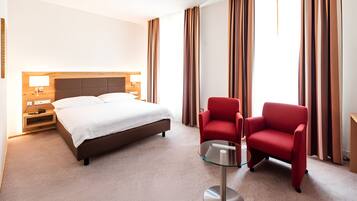 Una habitación de hotel con una cama, dos sillas rojas, una mesa de vidrio y una mesita de noche con lámpara.