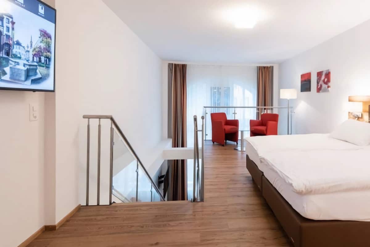 Quarto de hotel com uma cama grande, uma mesa com cadeira, TV na parede e vista para a cidade pela janela.