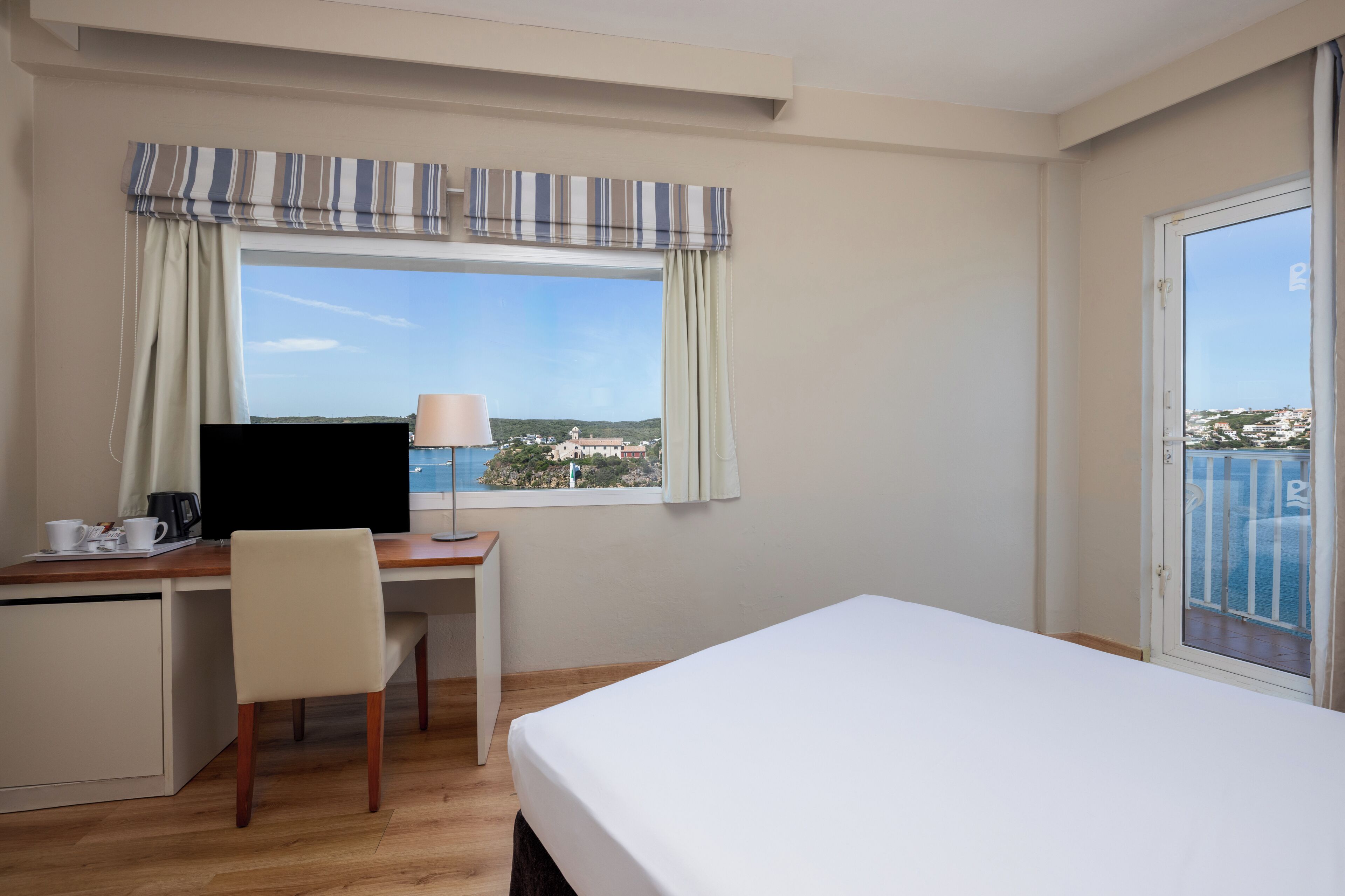 Quarto de hotel com cama, escrivaninha, cadeira e janela com cortinas.
