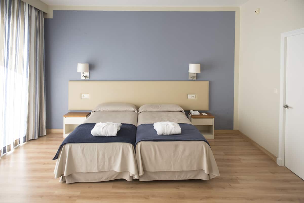 A hotel room with two beds, a wooden floor, a headboard with two wall-mounted lamps, and a door on the right.
