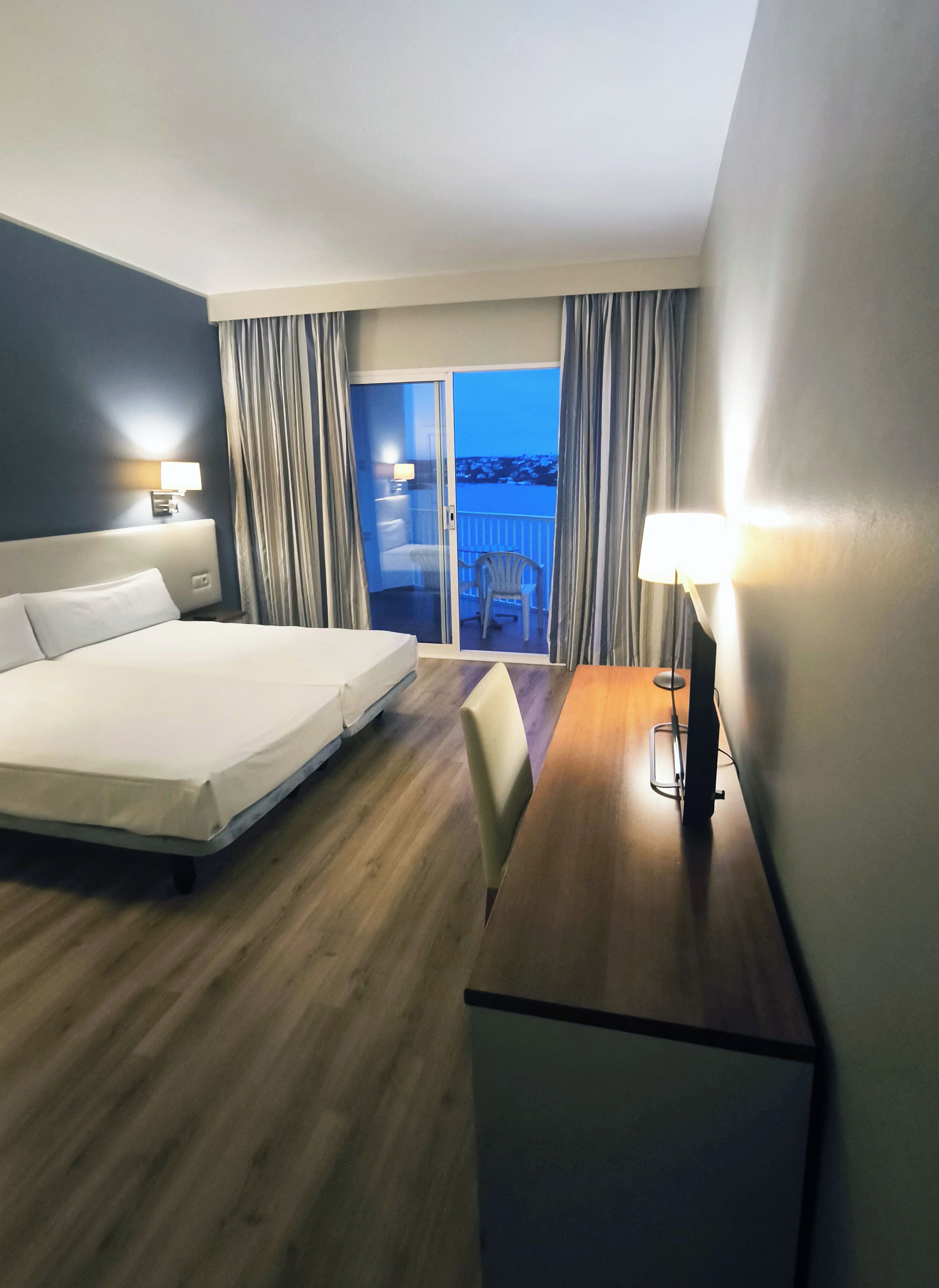 A hotel room with a bed, a desk, a chair, a lamp, a view of the outside, and curtains.