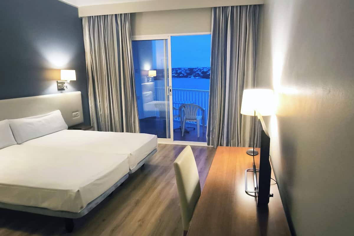 A hotel room with a bed, a desk, a chair, a lamp, a view of the outside, and curtains.