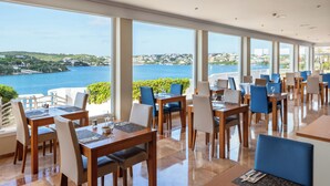 Un restaurante con varias mesas y sillas, amplias ventanas que ofrecen vistas al mar y las colinas, y un interior moderno y limpio.