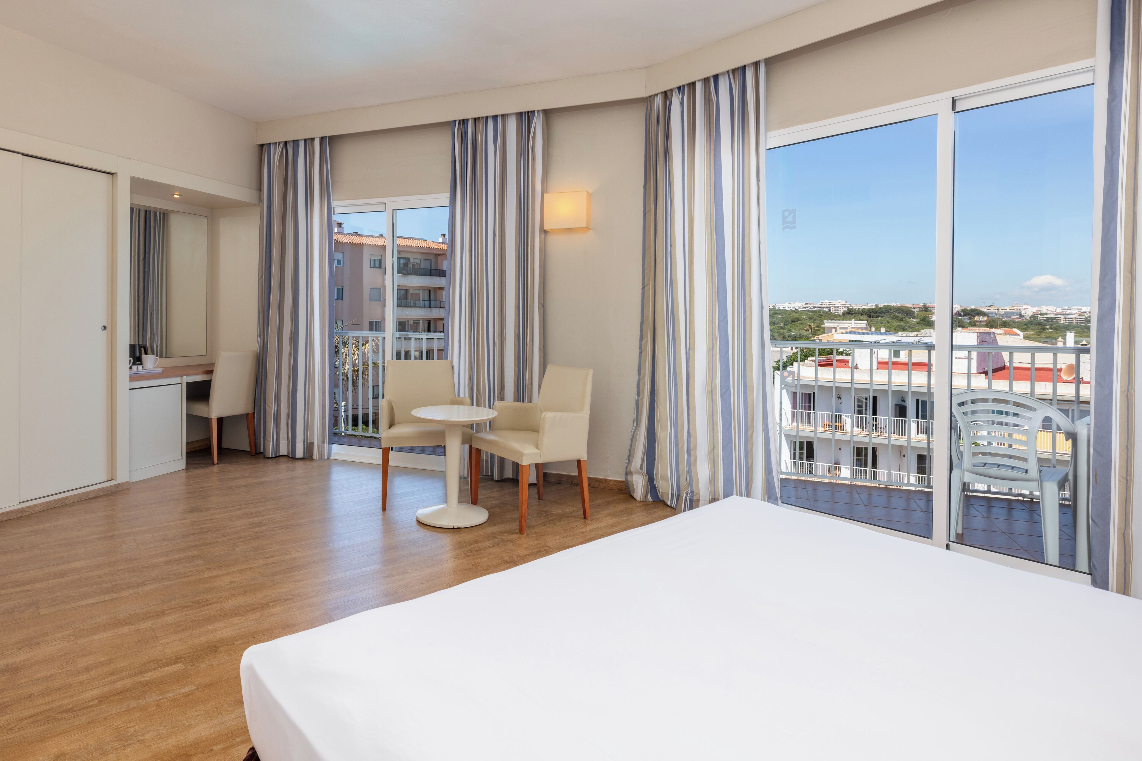 A hotel room with a bed, a desk, a chair, and a view of the cityscape through large windows.