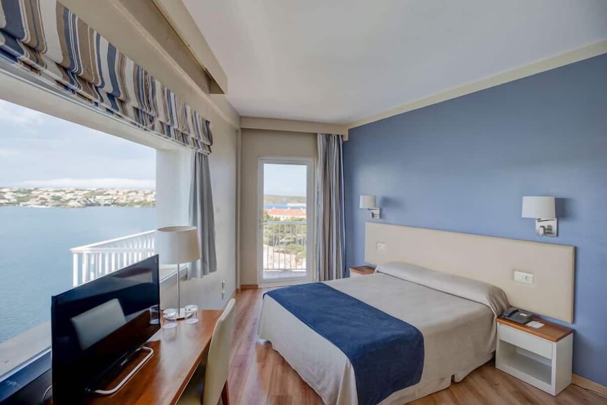 A hotel room with a bed, bedside table, TV, and a view of the sea.