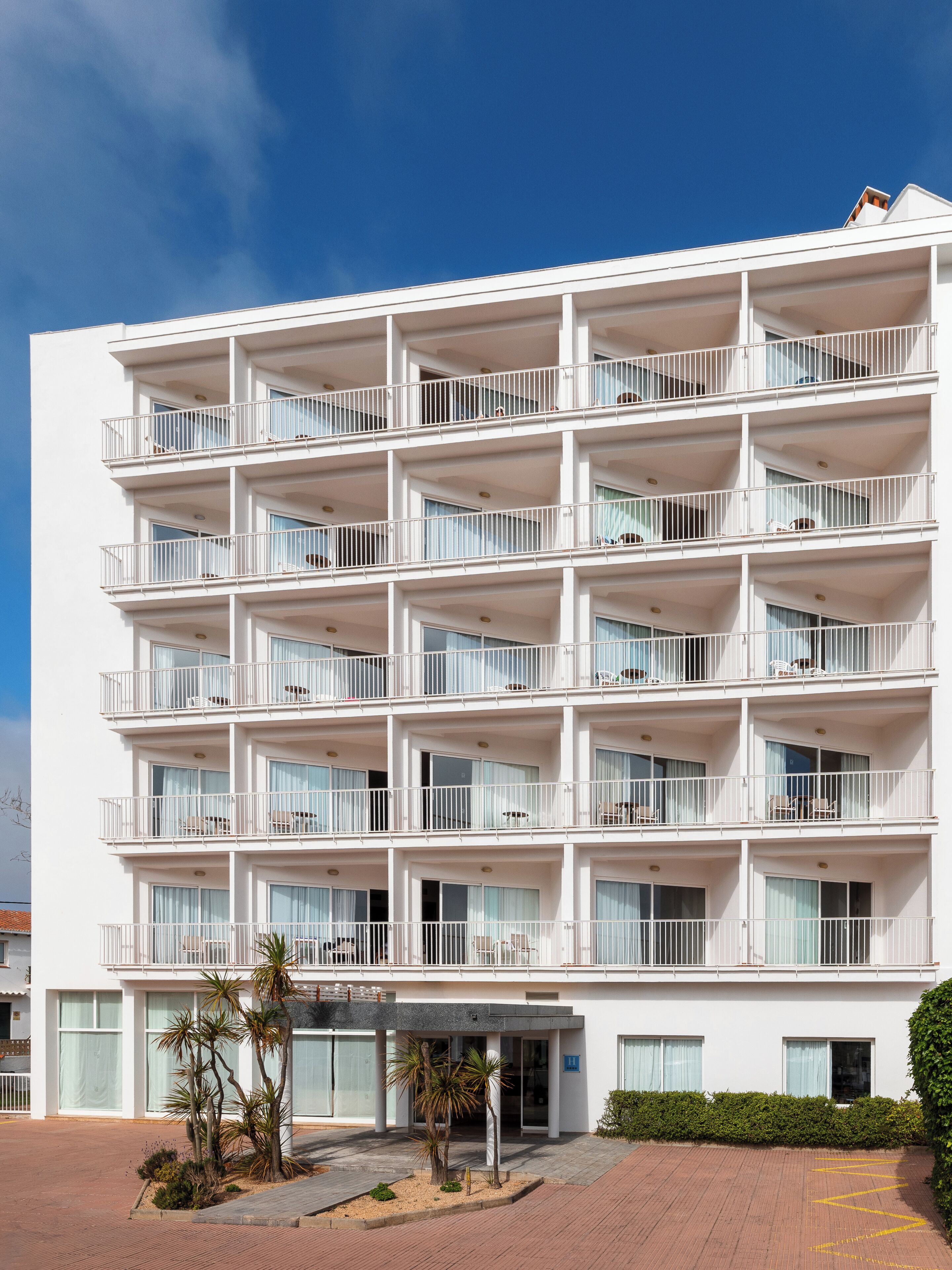 A multi-story hotel with balconies, a covered entrance, and a landscaped area with palm trees.