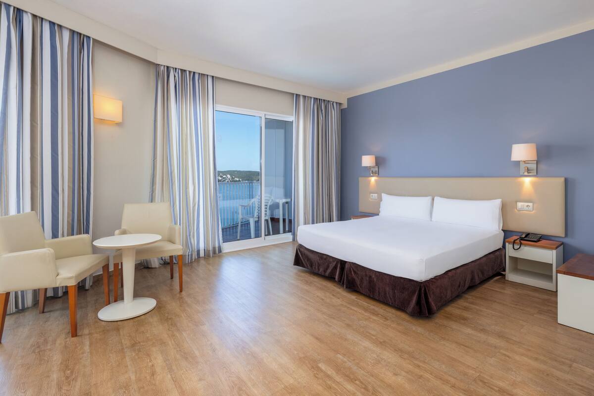 A hotel room with a bed, a small table, two chairs, a nightstand, and a view of the balcony.
