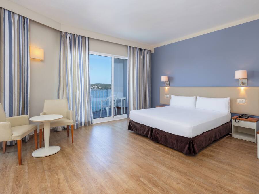 A hotel room with a bed, a small table, two chairs, a nightstand, and a view of the balcony.