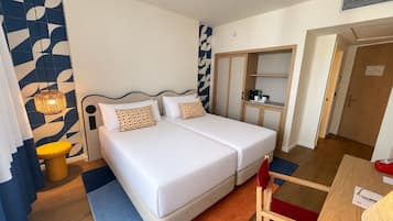 Cama doble con sábanas blancas, mesita de noche con una silla roja, armario de madera y una pequeña zona de kitchenette.