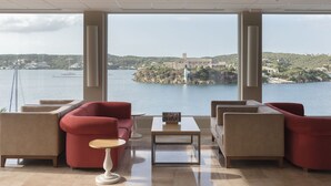 Una habitación moderna con un ventanal grande con vistas a una masa de agua, que incluye un sofá rojo, sillones color beige y una mesita.