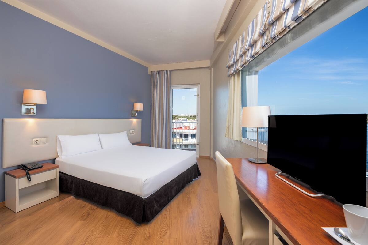 A hotel room with a bed, bedside tables, a desk with a chair, a television, and a window with a view.