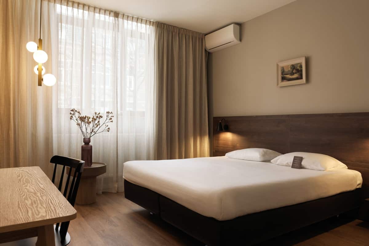 A hotel room with a bed, bedside table, chair, and a window with curtains.