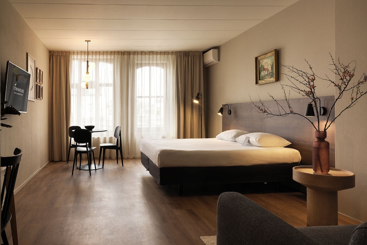 A hotel room with a large bed, a desk with two chairs, a TV, and a vase with branches.