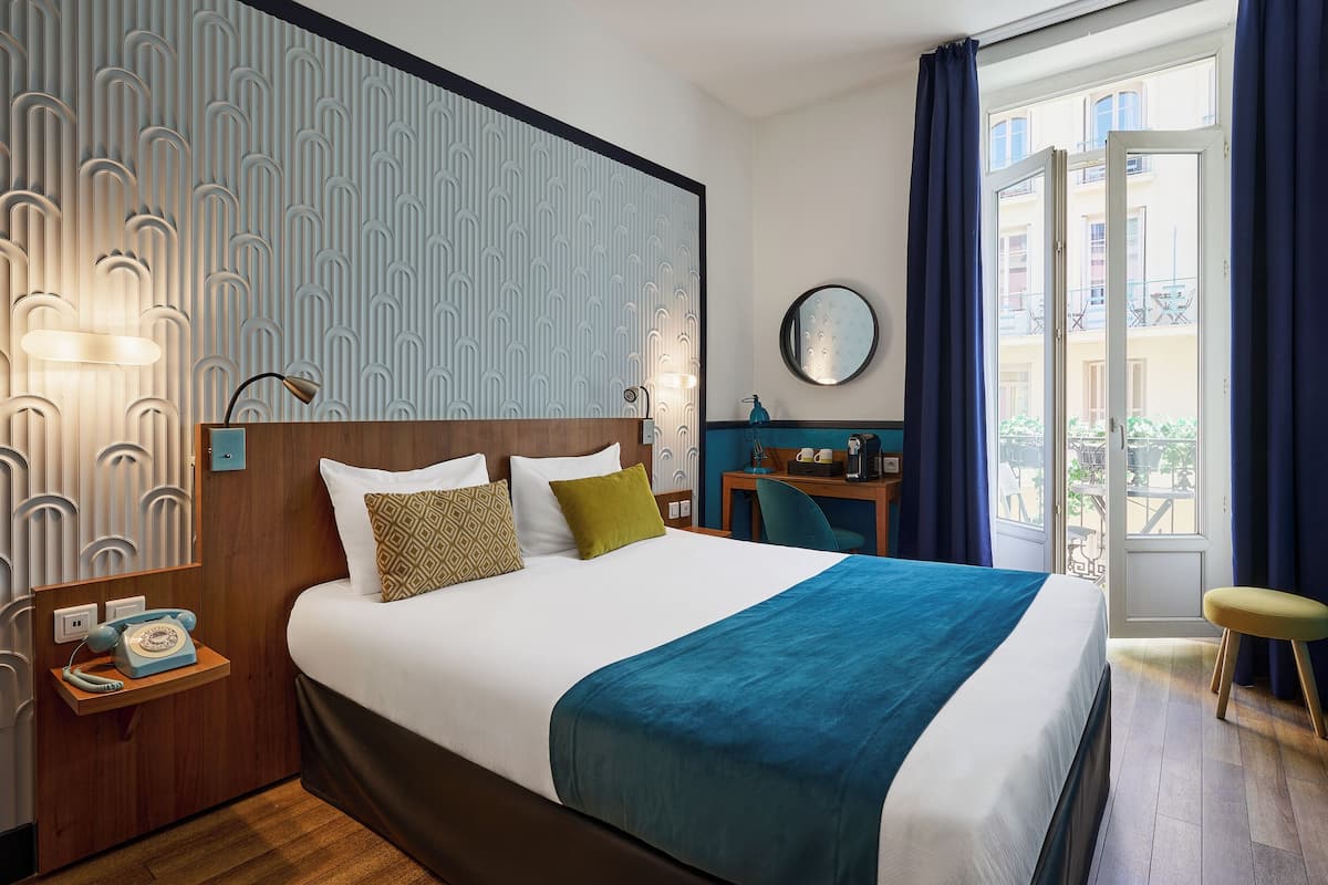 A hotel room with a bed, bedside table, lamp, and a view of the outside.