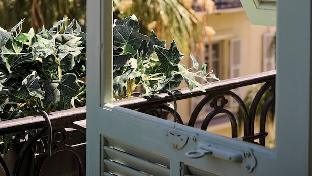 A window with a white frame and a partially open white shutter, overlooking a balcony with a metal railing and green foliage. - Hotel Nap By HappyCulture (Nice)