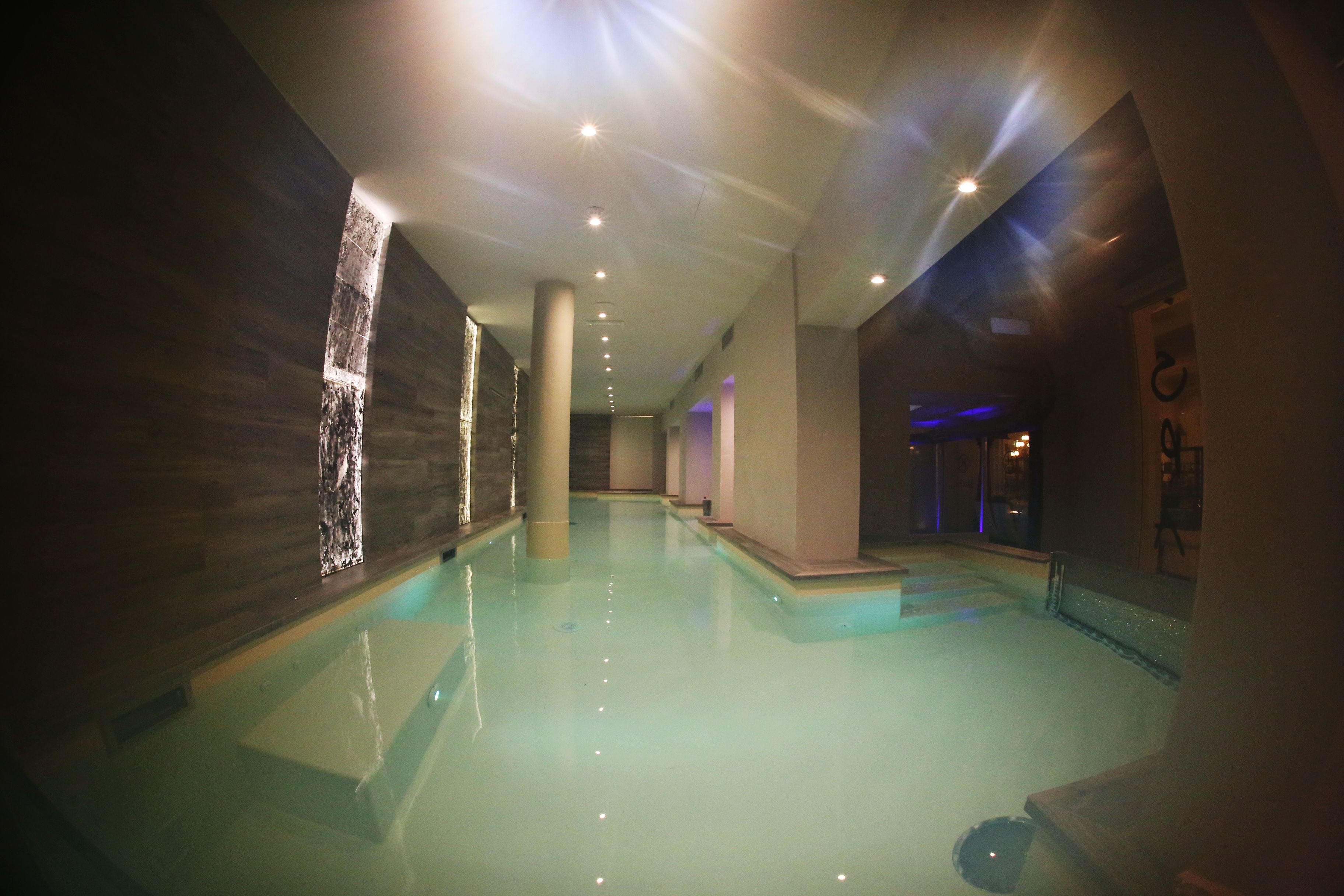 A spacious indoor swimming pool with a clear water surface, surrounded by walls with a textured finish and a series of evenly spaced lights on the ceiling.