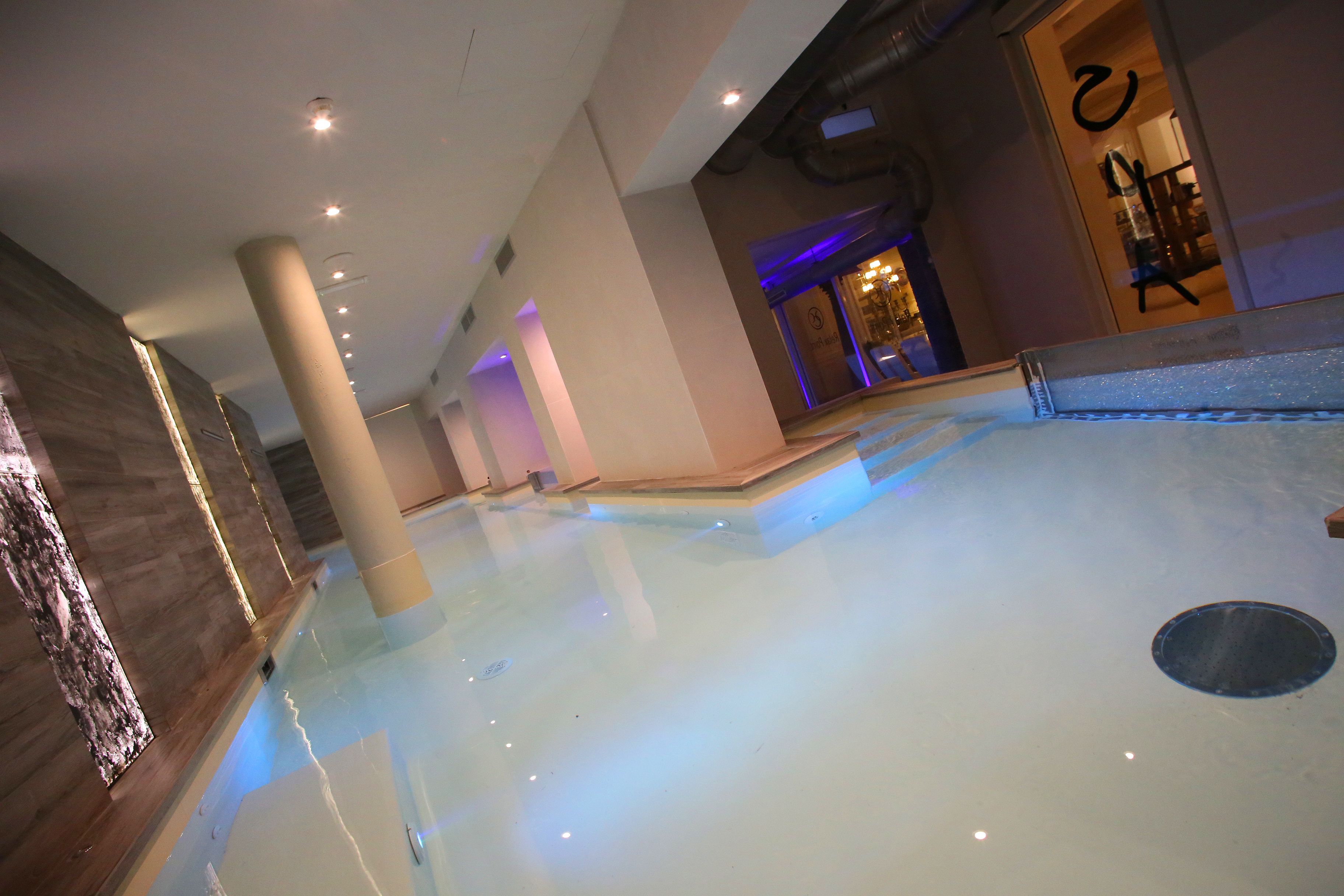 An indoor swimming pool with clear water, surrounded by beige walls and columns, and a visible entrance to another room.