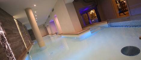 An indoor swimming pool with clear water, surrounded by beige walls and columns, and a visible entrance to another room.