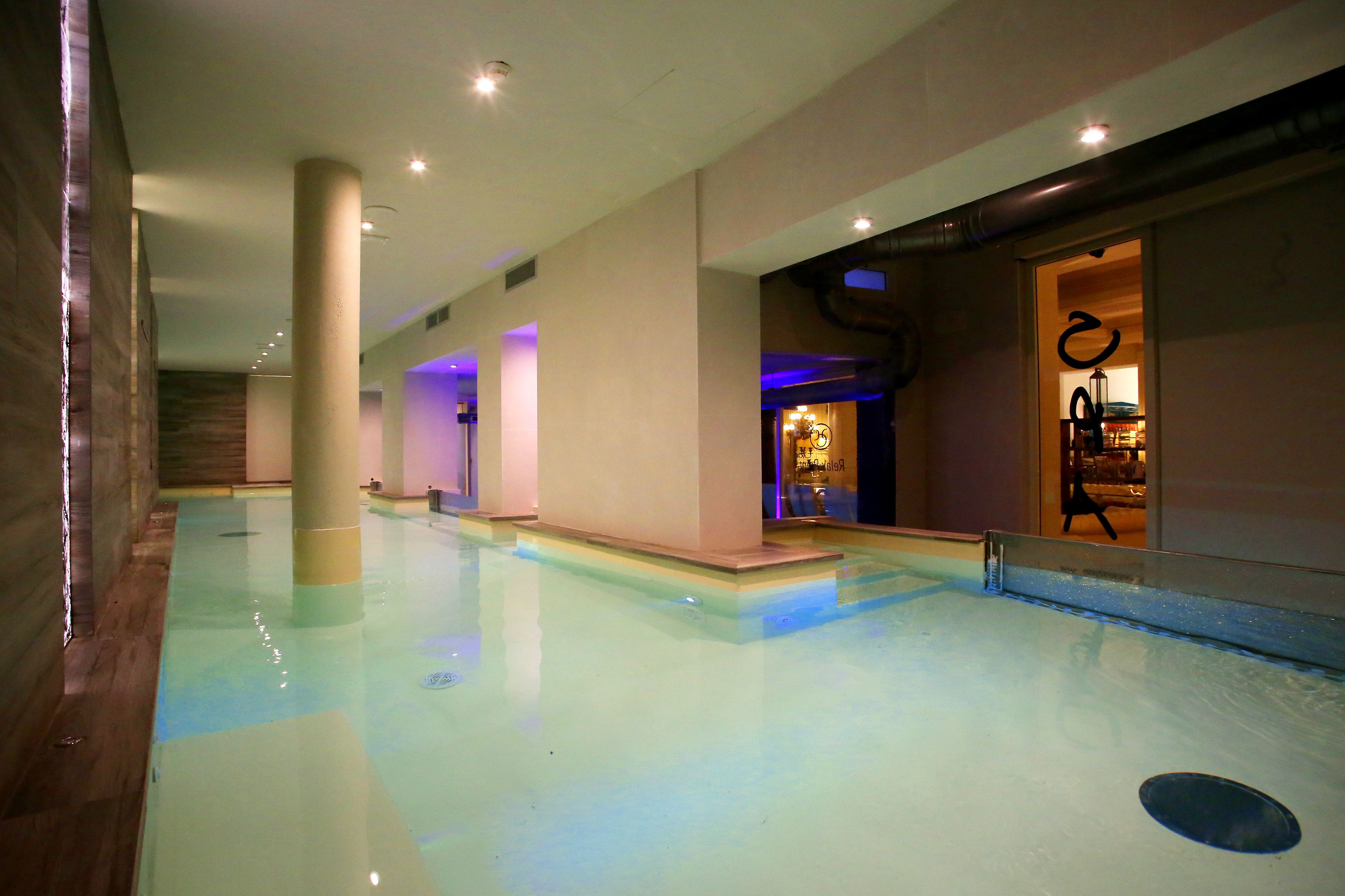 Una piscina interior amplia con agua cristalina, rodeada de paredes y columnas en tonos neutros, y una entrada visible a otra zona.