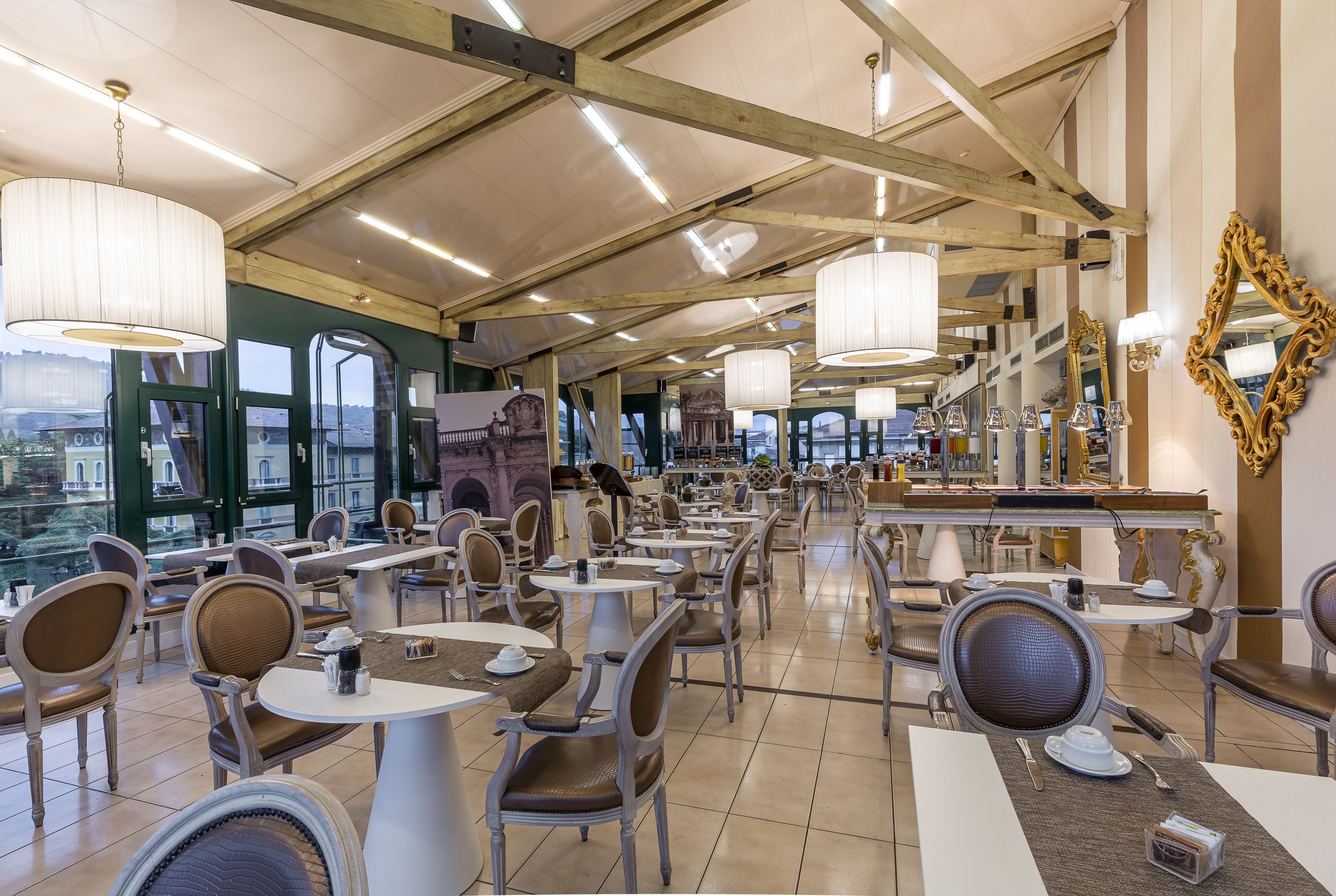 A spacious dining area with high ceilings, large windows, and elegant chandeliers. Tables and chairs are arranged neatly, and there are decorative mirrors on the walls.