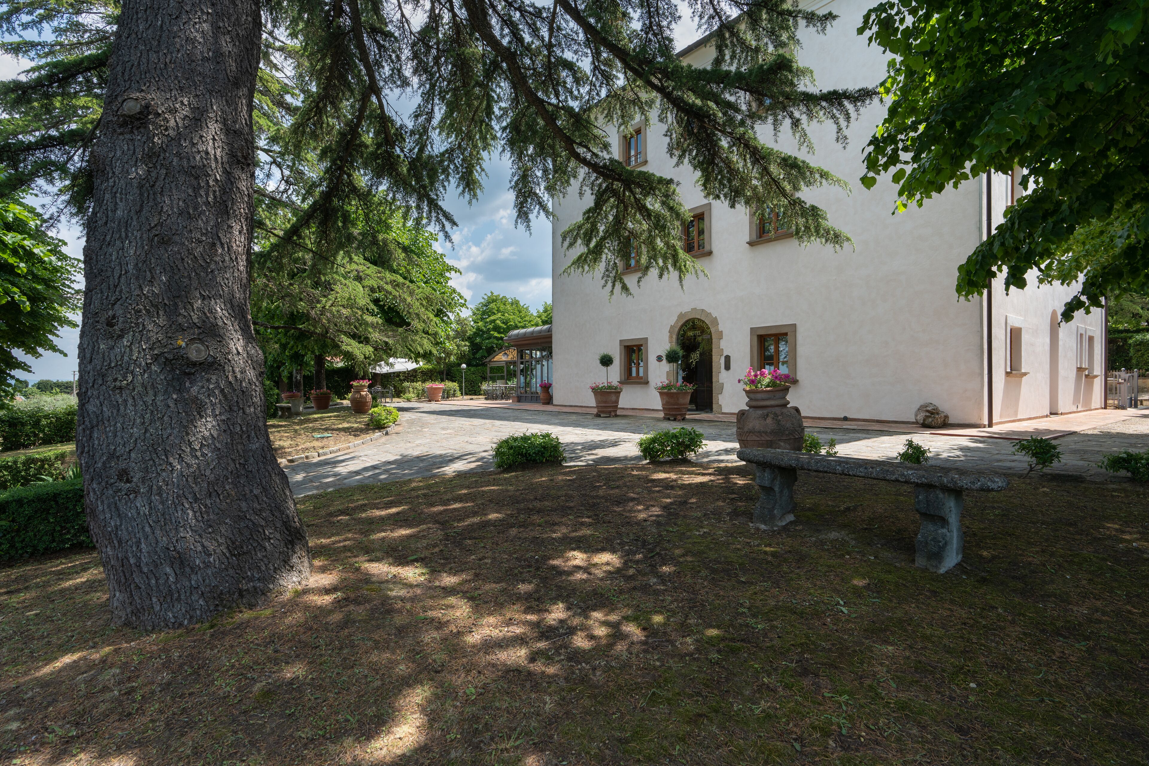 Un edificio de fachada blanca, un banco de piedra y un gran árbol en primer plano.