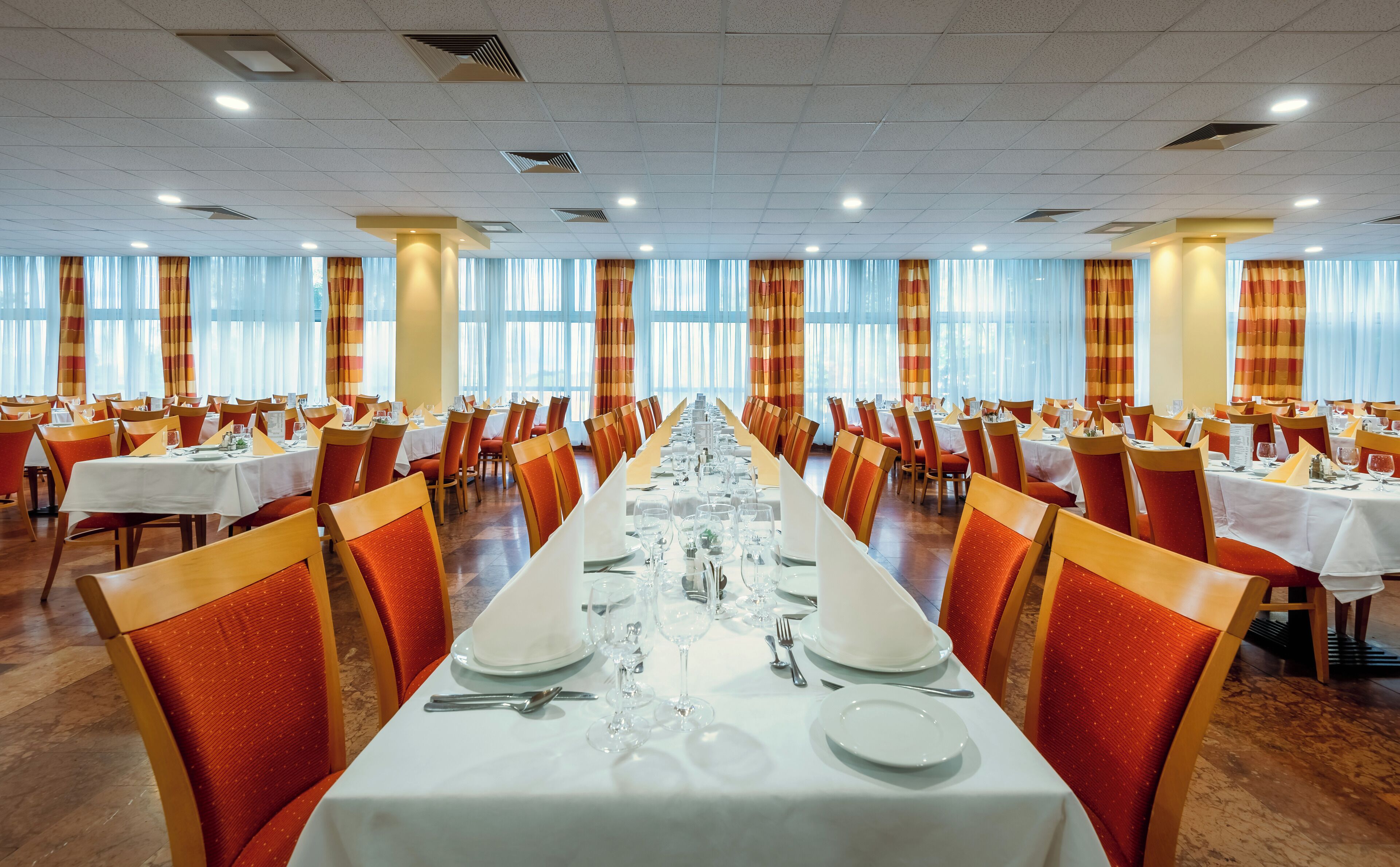 Une vaste salle de banquet, avec de longues rangées de tables dressées pour le repas, agrémentée de chaises orange et de nappes blanches.