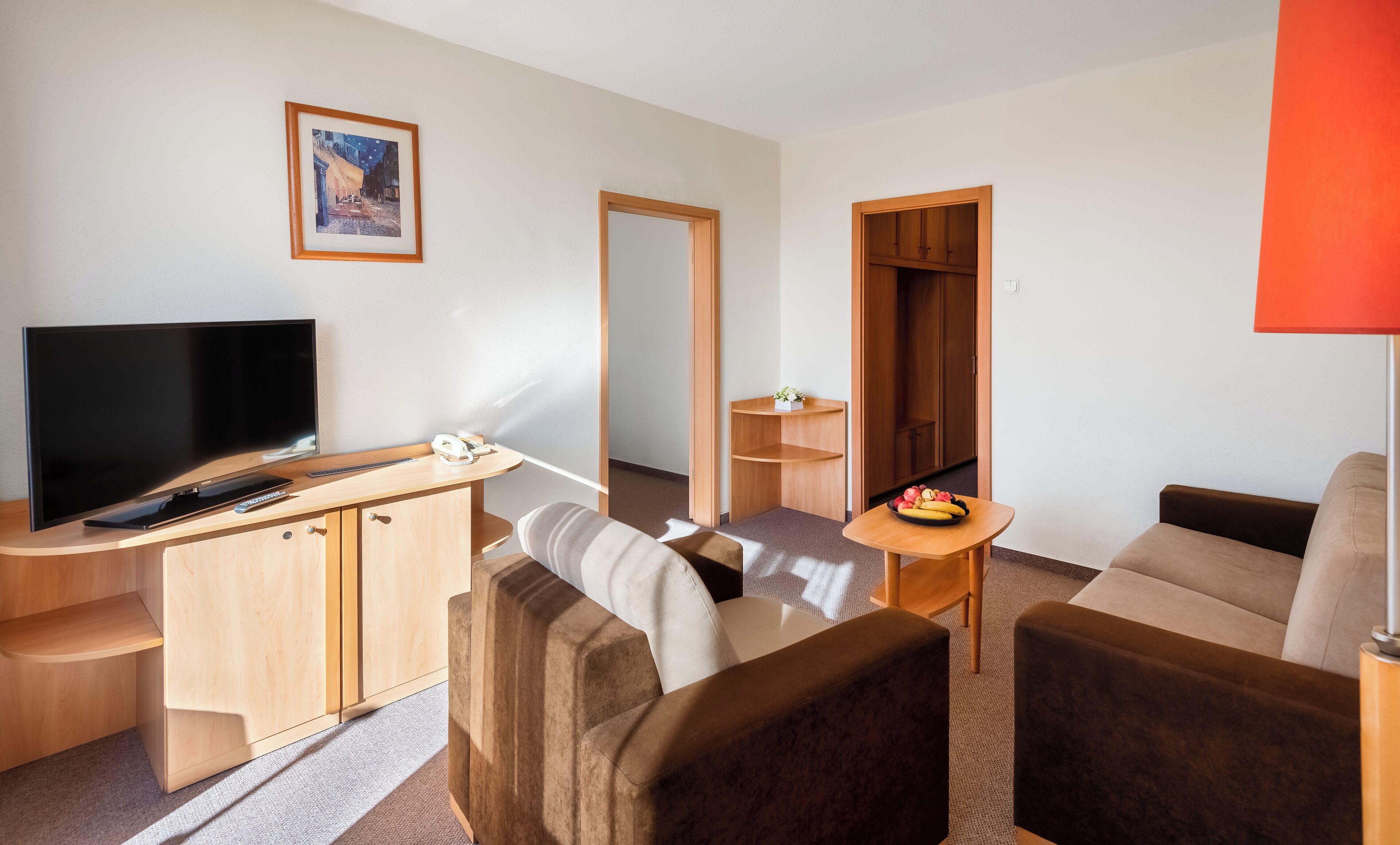 Une chambre d’hôtel moderne dotée d’une télévision à écran plat, de meubles en bois, d’une petite table avec des fruits, d’un canapé et d’un miroir.